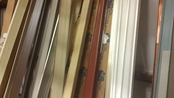 Various long and narrow wooden panels in beige, white, and red shades are stacked vertically in a cluttered workshop, conveying an atmosphere of busy organization.