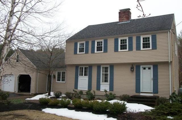 A two-story beige house with blue shutters and a red brick chimney. Snow patches on the ground, surrounded by bushes, evoke a winter feel.