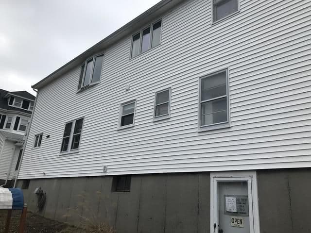 White multi-story building with several windows on a cloudy day. A sign reading "Open" is on a door at the concrete lower level. Nearby buildings visible.