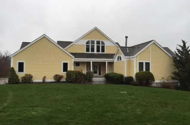 A yellow house with multiple gables and white trim sits amid a lush green lawn. It has a welcoming entryway and a cloudy sky backdrop, creating a serene mood.