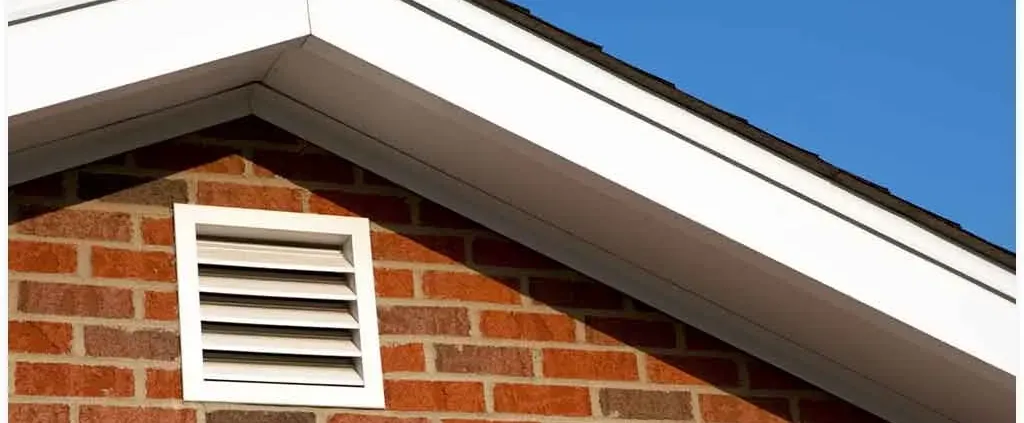 Gable roof with white trim on a red brick house, featuring a white rectangular vent, set against a clear blue sky. The scene is bright and clean.