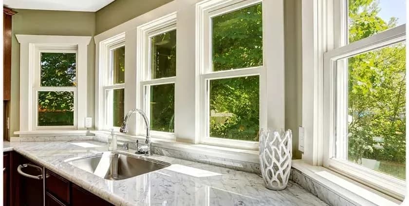 A bright kitchen with white-framed windows showcasing green foliage outside. A sleek marble countertop features a stainless steel sink and a vase.