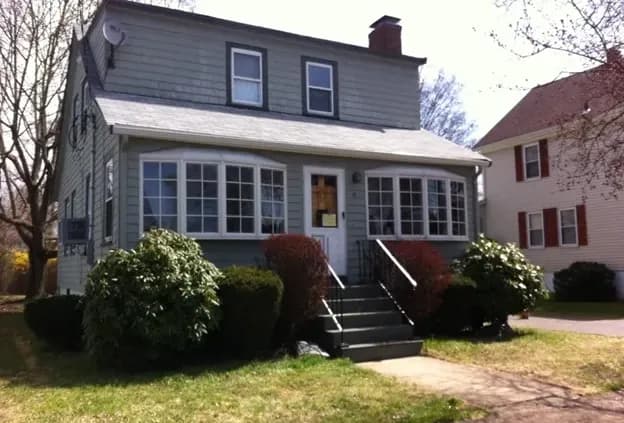 A two-story gray house with a front porch, large windows, and a chimney. Bushes and a tree surround the walkway, creating a welcoming atmosphere.
