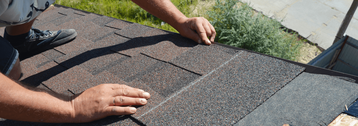 Person installing asphalt shingles on a roof, hands pressing down firmly. The scene conveys precision and focus, with greenery in the background.