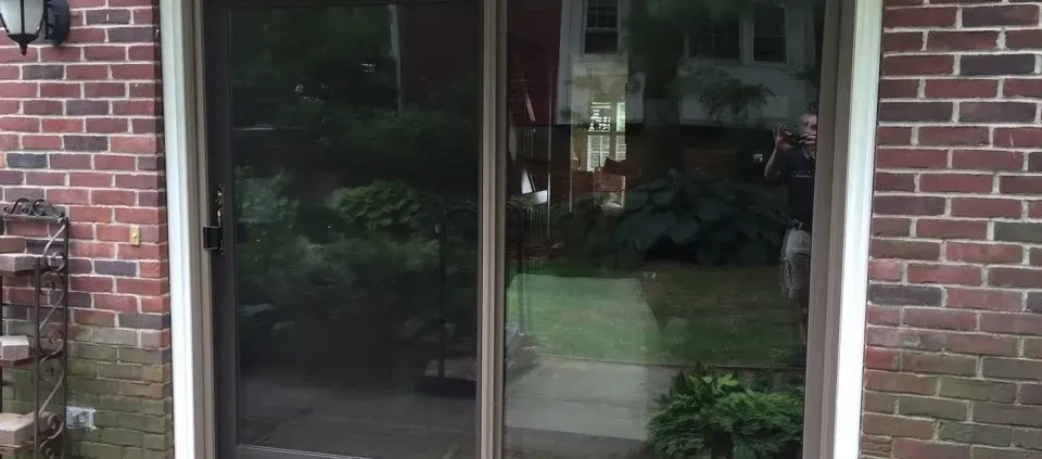 A glass sliding door set in a brick wall reflects greenery from a garden, with a glimpse of ferns and potted plants nearby, creating a serene outdoor atmosphere.