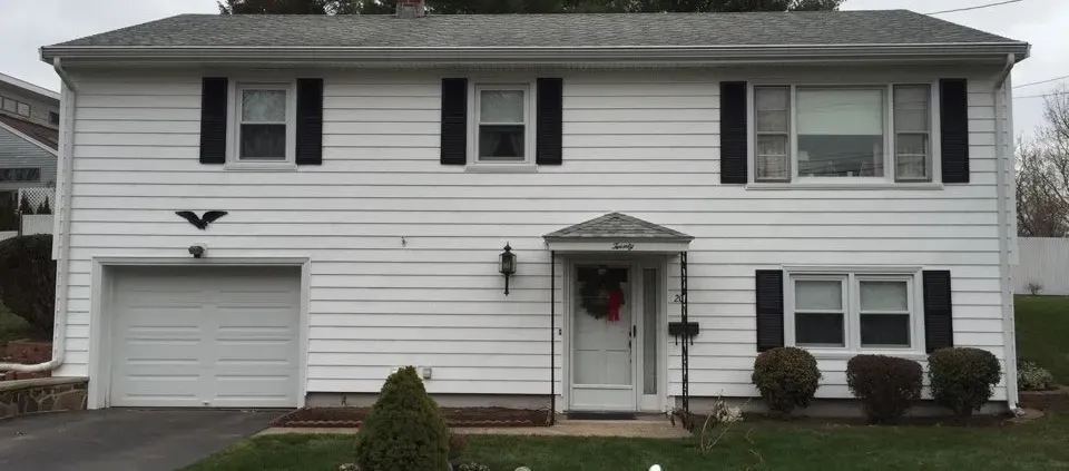 A white split-level house with dark shutters, a central front door, and a single-car garage. Neatly trimmed bushes line the front walkway.