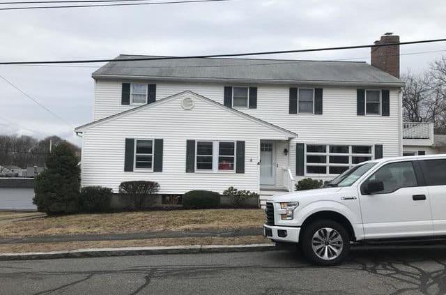 A two-story white house with black shutters and a chimney. A white pickup truck is parked in front. Overcast sky gives a subdued, neutral tone.