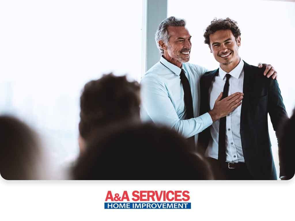 Two smiling men in suits share a friendly gesture at an indoor event, conveying camaraderie. The text "A&A Services Home Improvement" is visible below.