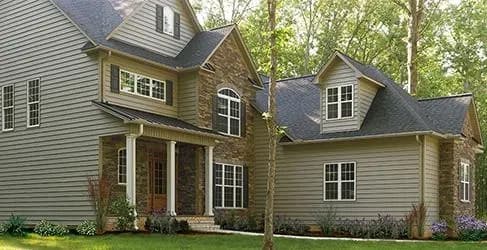 A two-story suburban house with gray siding and stone accents set in a lush, green garden. Tall trees surround the peaceful, inviting home.