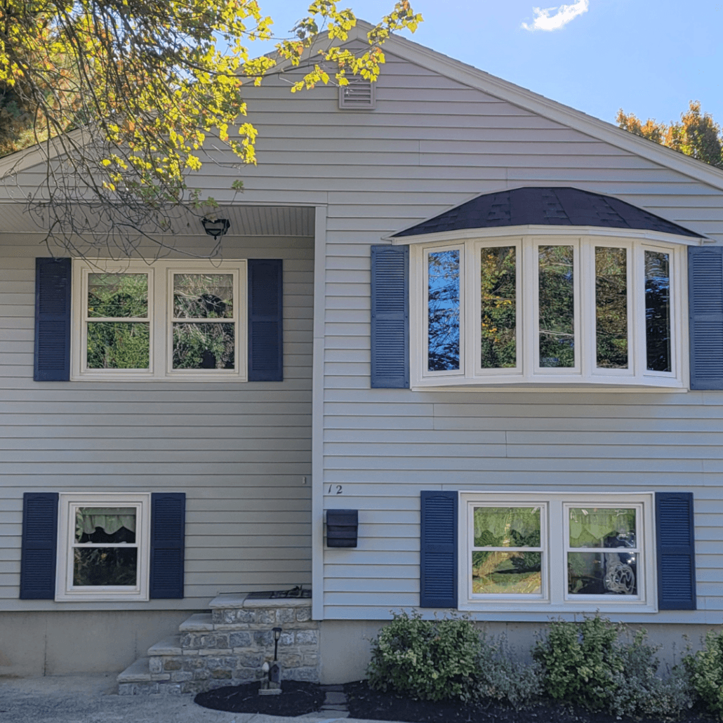 A two-story house with light gray siding and blue shutters. A bay window and neat shrubs add charm. A tree with autumn leaves shades the scene.