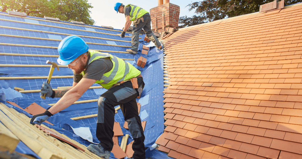 Two construction workers in safety gear repair a roof under clear skies, using hammers and new shingles. The scene conveys focus and teamwork.