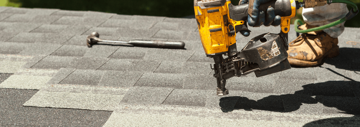Worker uses a yellow nail gun to install asphalt shingles on a roof. A hammer lies nearby. The scene conveys focus and craftsmanship.