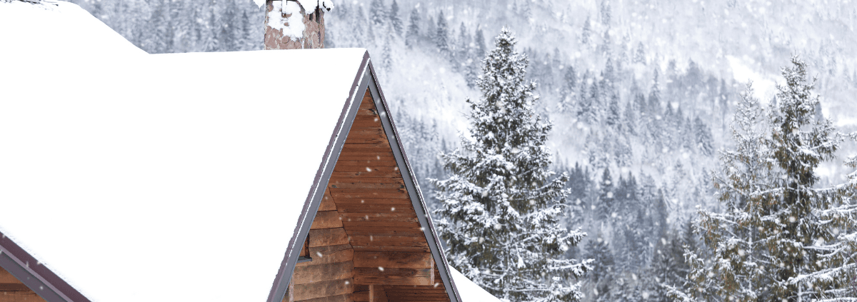 Snow-covered wooden cabin roof with a backdrop of frosty pine trees and misty, snow-laden mountains, conveying a serene winter atmosphere.