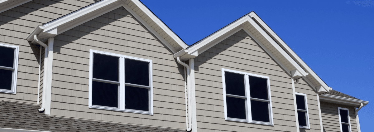 Close-up of a beige house exterior featuring two sets of double windows beneath steep gables, against a clear blue sky, conveying a calm and sunny atmosphere.