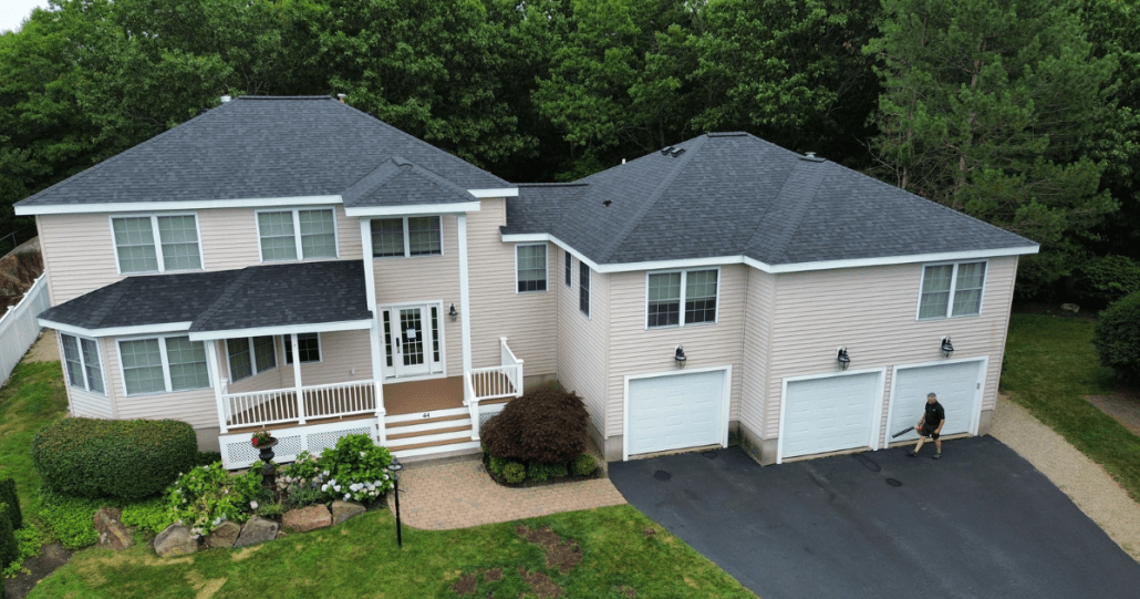 Two-story beige house with dark blue roof, surrounded by lush greenery. Two-car garage, landscaped garden, and person sitting on driveway edge.