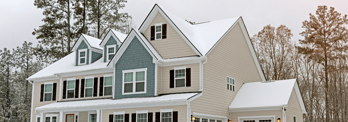 A two-story house with a snow-covered roof and modern design is set against a backdrop of tall trees under an overcast sky, conveying a serene winter feel.