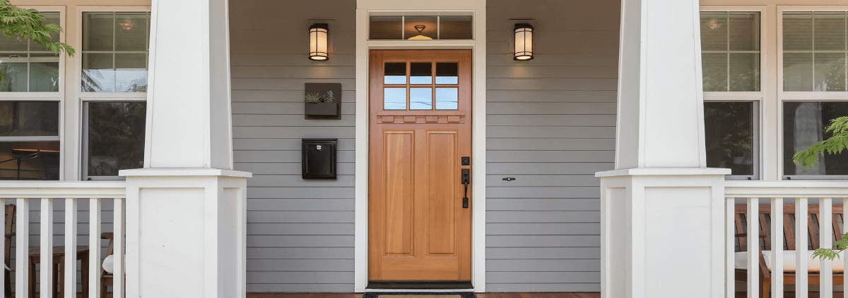A welcoming front porch with a wooden door, flanked by white columns and lanterns. The gray siding and wooden floor create a warm, inviting ambiance.
