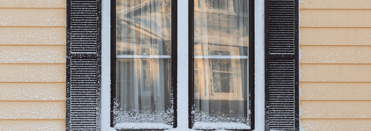 A snow-dusted window on a yellow house, with dark shutters framing icy panes. Reflective of a serene winter morning, the scene is calm and quiet.