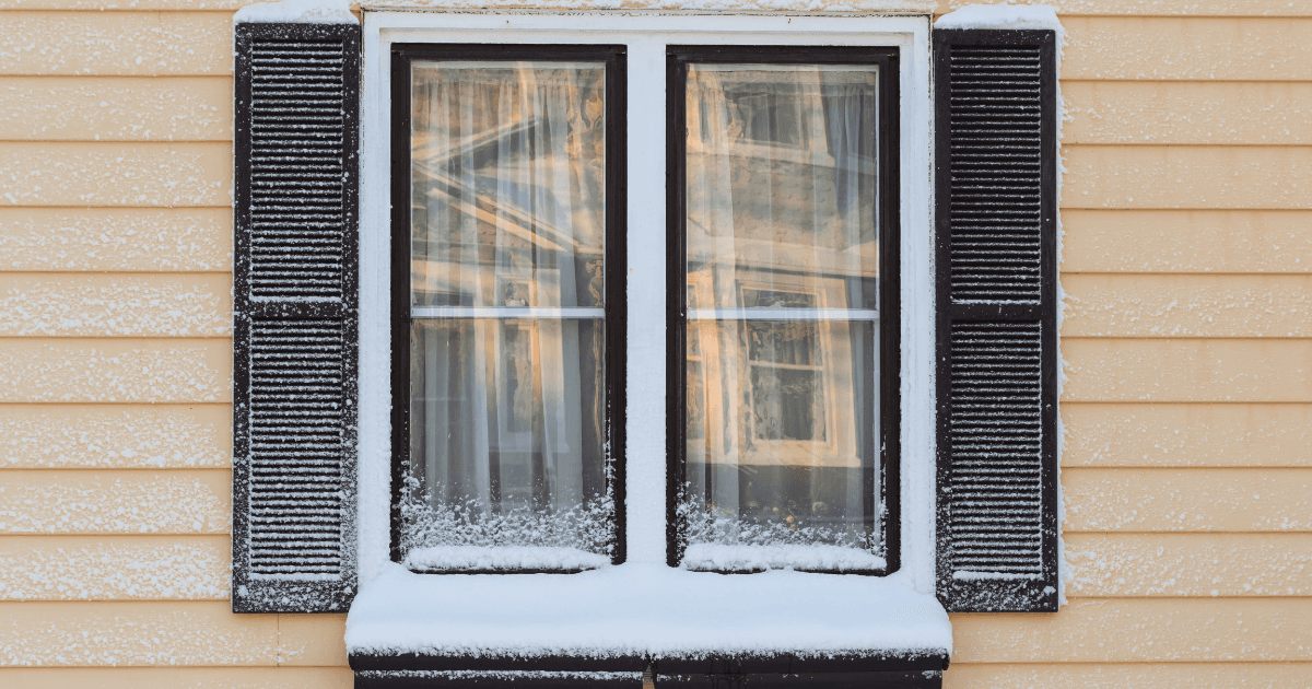 A snowy window with black shutters on a yellow house, covered in a dusting of snow, creating a cozy, wintertime atmosphere.