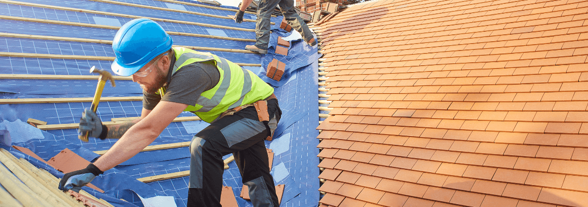 Construction workers installing shingles on a roof. A man in a blue helmet and yellow vest hammers nails, while another worker prepares materials.