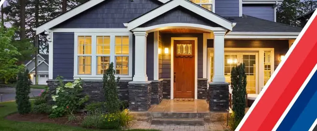 Charming house with a lit porch at dusk featuring navy siding, white trim, and stone accents. Manicured lawn and vibrant garden in front. Cozy ambiance.