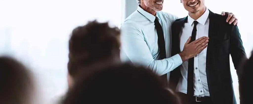 Two men in business attire share a friendly moment in an office, with one man smiling and patting the other's chest. The audience is slightly blurred in the foreground.