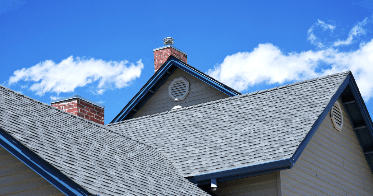 Gabled house roof with gray shingles against a bright blue sky and scattered clouds. Two red brick chimneys add contrast and texture to the scene.