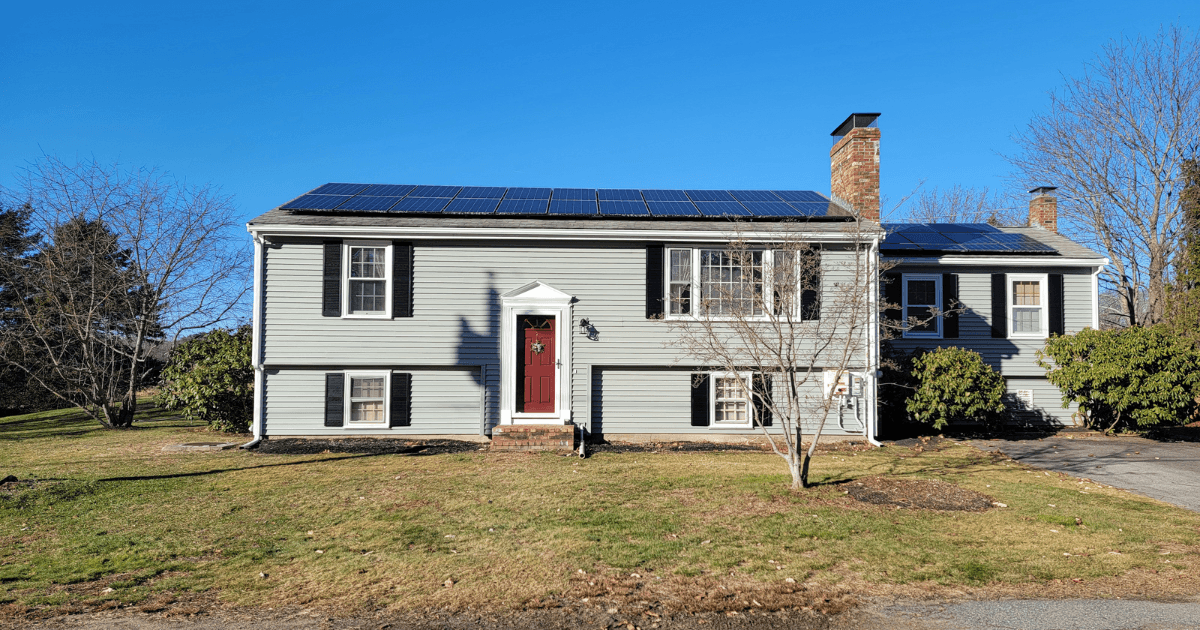 A gray split-level house with solar panels on the roof, a red front door, and black shutters. It's surrounded by a lawn and leafless trees under a clear blue sky.
