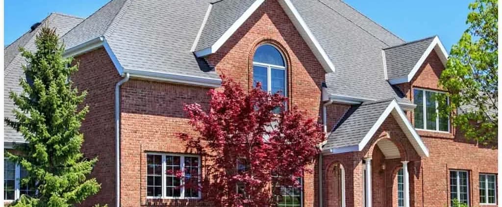 A large, elegant brick house with a steep, gray roof is surrounded by trees. The front features a prominent arched window and a vibrant red tree.