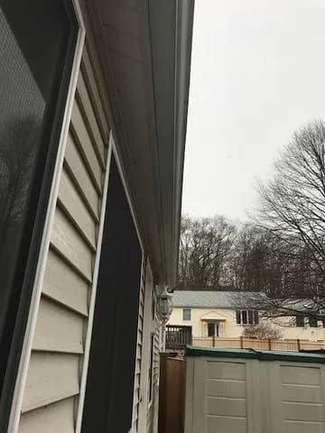 Side view of a beige house with a black window screen, next to a green-roofed shed. Background features leafless trees and cloudy sky. Quiet suburban setting.