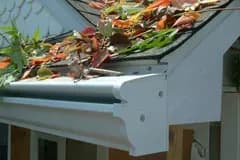 A house gutter is clogged with colorful autumn leaves and debris, highlighting maintenance needs. The gutter is attached to a sloping roof.