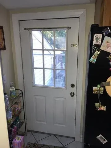 A white door with a windowpane grid has sunlight streaming through, illuminating a cozy kitchen with a black fridge covered in magnets and drawings.