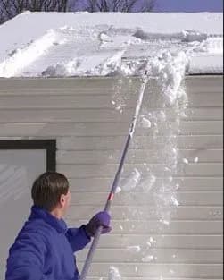 A person in a purple jacket uses a roof rake to clear thick snow from a white roof, with snow mid-fall, under a clear blue sky.