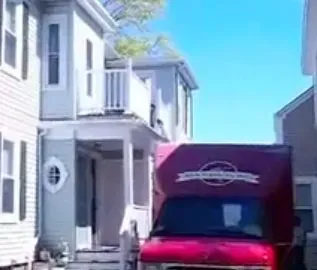 A red delivery truck parked in front of a light gray townhouse on a sunny day. The truck partially blocks the view of the house's front porch.