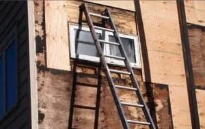 A metal ladder leans against a partially boarded-up wall of a house, reaching a small window. The scene suggests renovation or repair work in progress.
