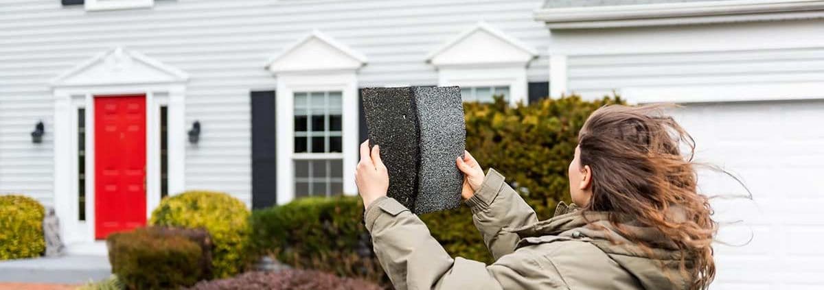 A person with long hair holds two roofing shingles while facing a gray house with a bright red door and white trim, conveying a sense of evaluation.