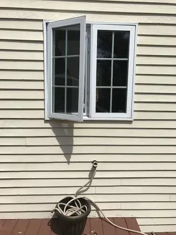 An open window on a beige siding wall casts a shadow. Below, a garden hose is coiled in a black pot on a brown deck. The scene is bright and sunny.