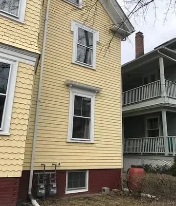 A yellow house with white trim, featuring vertical windows and a brick foundation. Nearby, a red rain barrel and an adjacent gray house are visible.