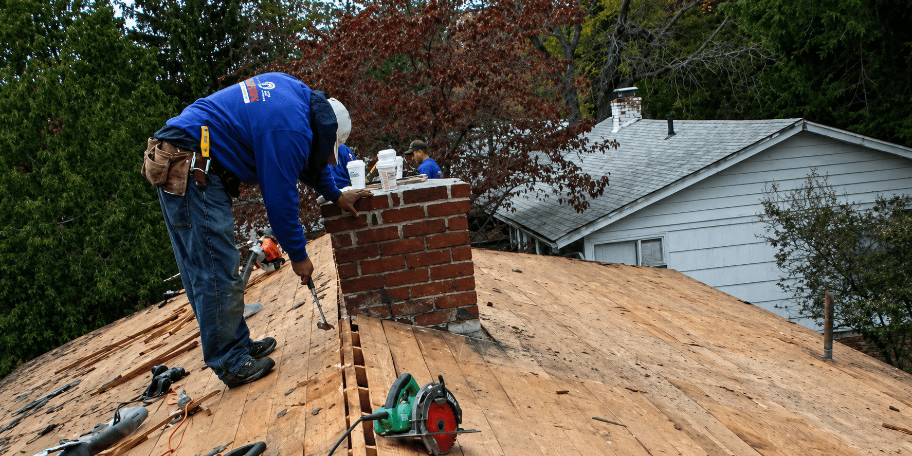 A construction worker in a blue shirt is repairing a wooden roof near a brick chimney. Tools and equipment are scattered around, with a house and trees in the background.