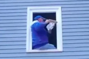 A person in a blue jacket and hat kneels in a tall, narrow window of a blue-paneled building, adjusting something with a cloth.