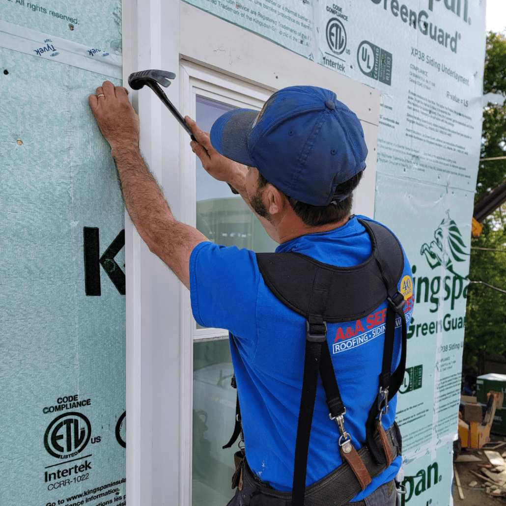 A worker in a blue shirt and cap uses a hammer to install siding on a house under construction. He wears a safety harness, conveying focus and diligence.