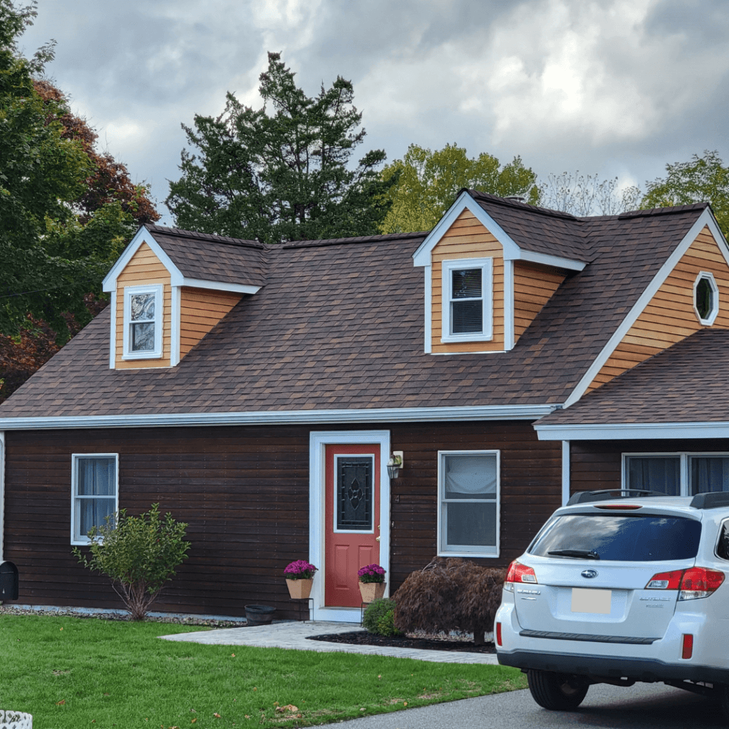 A charming suburban house with brown siding, white trim, and two dormer windows. Red front door, white SUV in driveway, cloudy sky overhead.