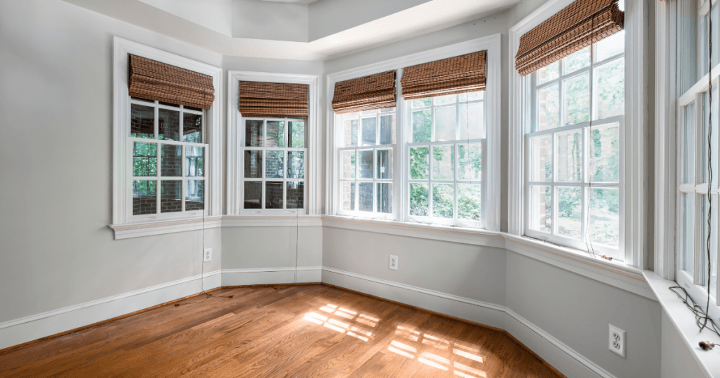 A sunlit, empty room with light gray walls and hardwood floors. Five large windows with brown wicker shades offer views of lush greenery outside.