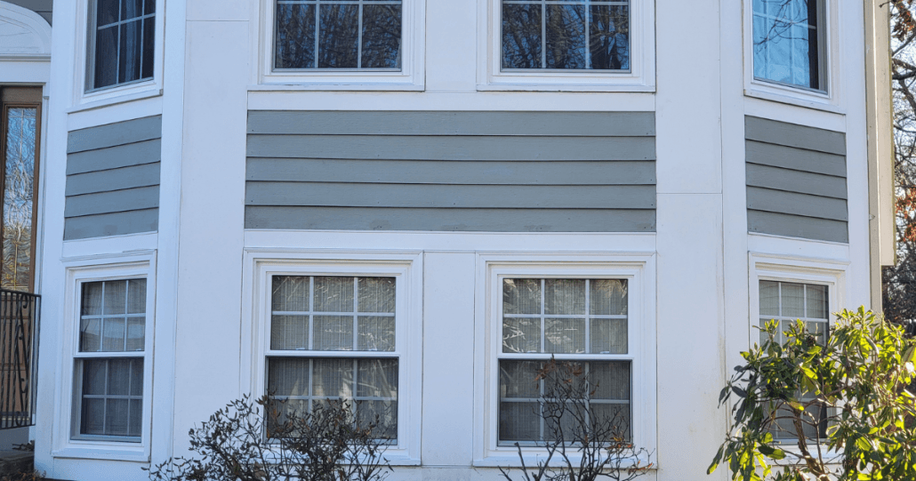 A house exterior with light gray and white siding features a bay window with six rectangular grid windows. Sparse bushes are in the foreground.
