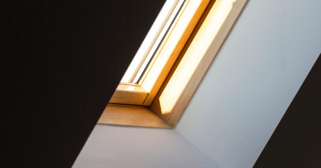 A sunlit wooden-framed skylight window in a slanted ceiling, casting soft shadows and creating a warm, serene ambiance in the room.