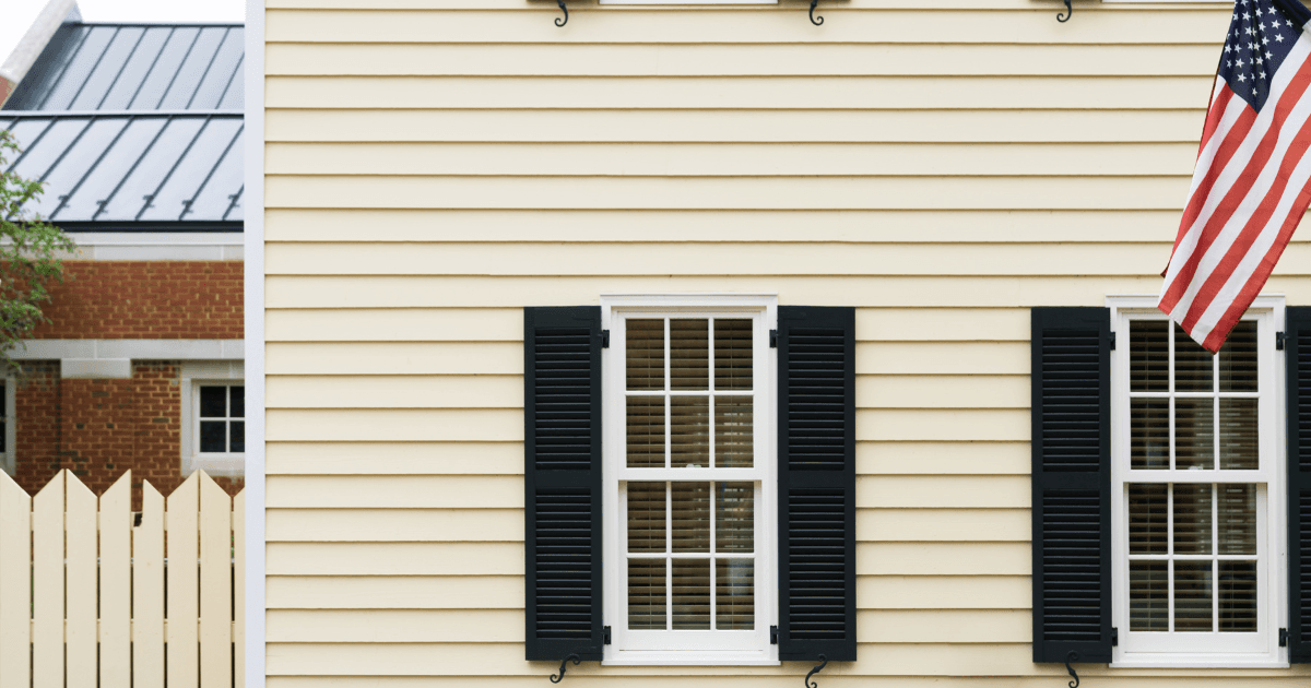 A beige house exterior with two white-paned windows framed by black shutters. An American flag hangs on the right. Fenced yard and brick building in the background.