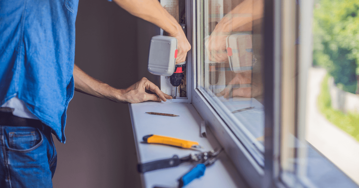 A person in a blue shirt uses an electric drill to fix a window frame. Nearby, a utility knife and pliers rest on the windowsill. Bright, focused mood.