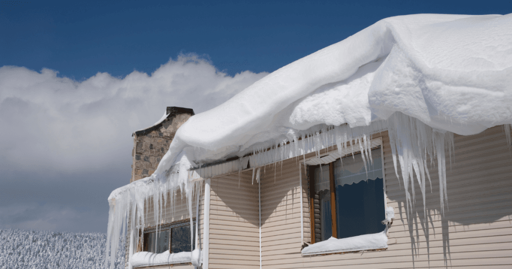 A house roof covered in thick snow and icicles against a bright blue sky. The scene conveys a cold, wintery atmosphere with clear weather.