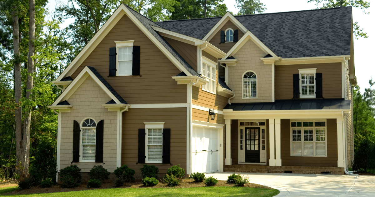 Two-story suburban house with tan siding, white accents, and black shutters. It has a gable roof, arched windows, and a two-car garage, surrounded by trees.