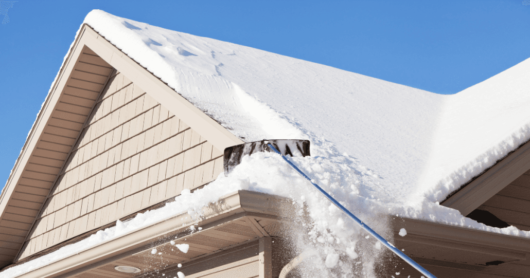 A roof rake removes snow from a sloped roof of a house, set against a clear blue sky. The scene conveys a chilly, winter maintenance task.